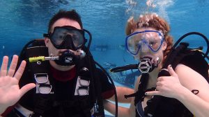 man waving and woman signing underwater in scuba gear