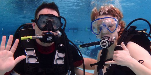 man waving and woman signing underwater in scuba gear